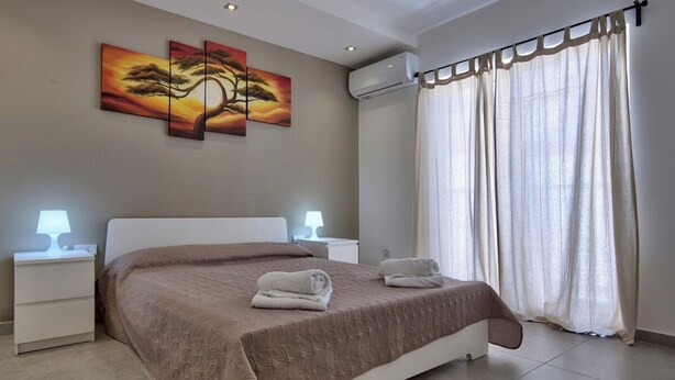 A bedroom with a large bed, two bedside tables with lamps, a wall-mounted air conditioner, and a window with curtains. - Off the Strand Gzira 3-bedroom Apartment (Gzira)