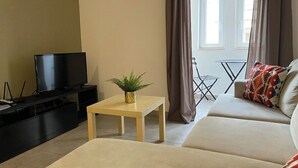 A living room with a TV, a small table with a plant, a sofa, and a window with curtains. - Off the Strand Gzira 3-bedroom Apartment (Gzira)
