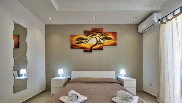 A hotel room with a large bed, two bedside tables with lamps, a wall-mounted air conditioner, and a painting of a tree. - Amazing Airy Apartment Surrounded by All Amenities (Gzira)