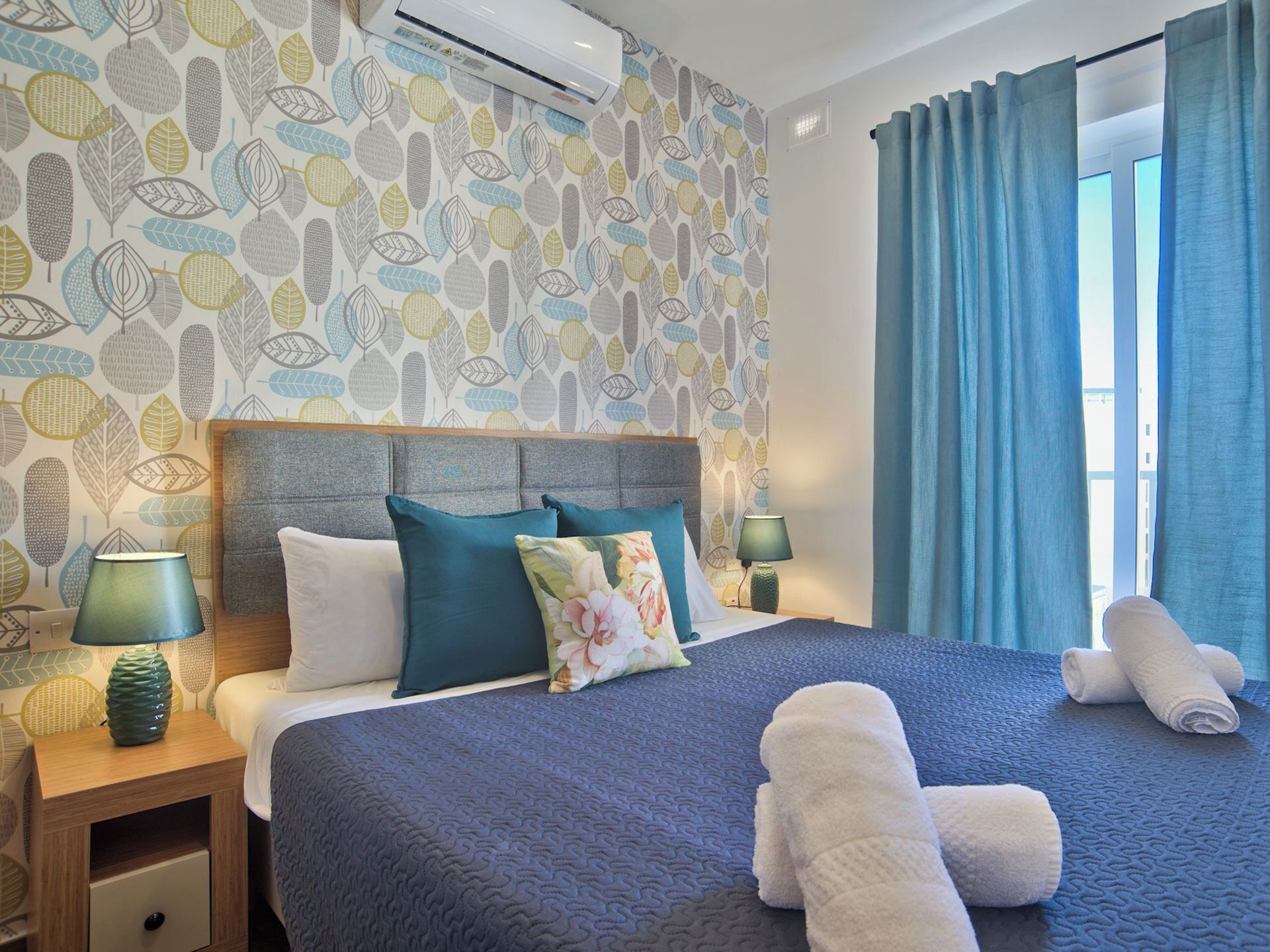A hotel room with a patterned wallpaper, a bed with teal and white bedding, a headboard, two bedside lamps, a nightstand, and a window with blue curtains.
