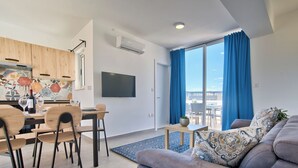 Uma sala de estar moderna com um sofá cinzento, uma área de jantar com cadeiras de madeira, uma TV fixada na parede e uma janela grande com cortinas azuis.