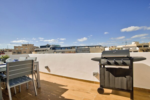 A rooftop patio with a barbecue grill, a table with chairs, and a view of surrounding buildings. - Amazing Duplex Penthouse With Jacuzzi and BBQ (Sliema)