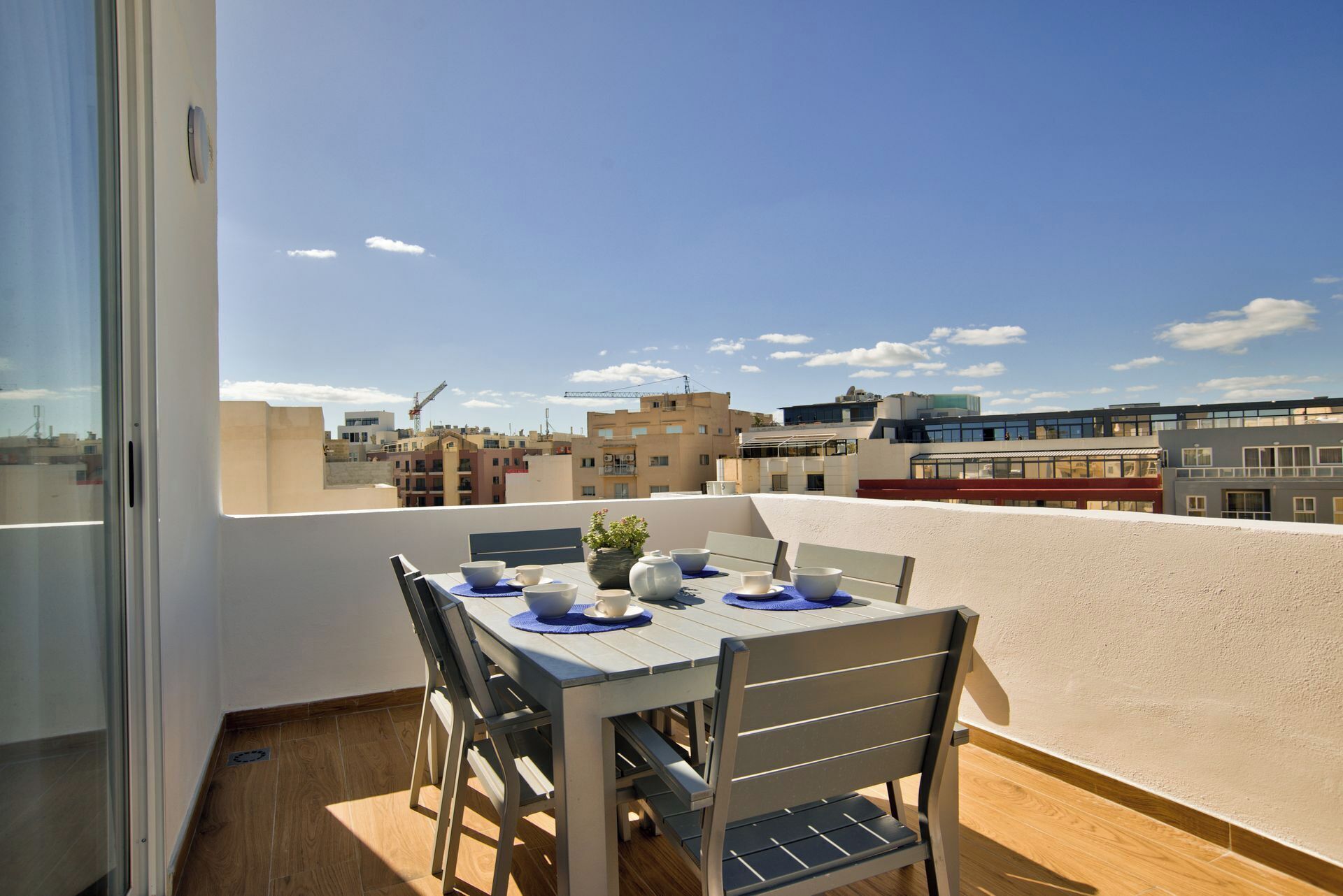 A balcony with a table set for two, overlooking a cityscape with buildings and construction cranes.