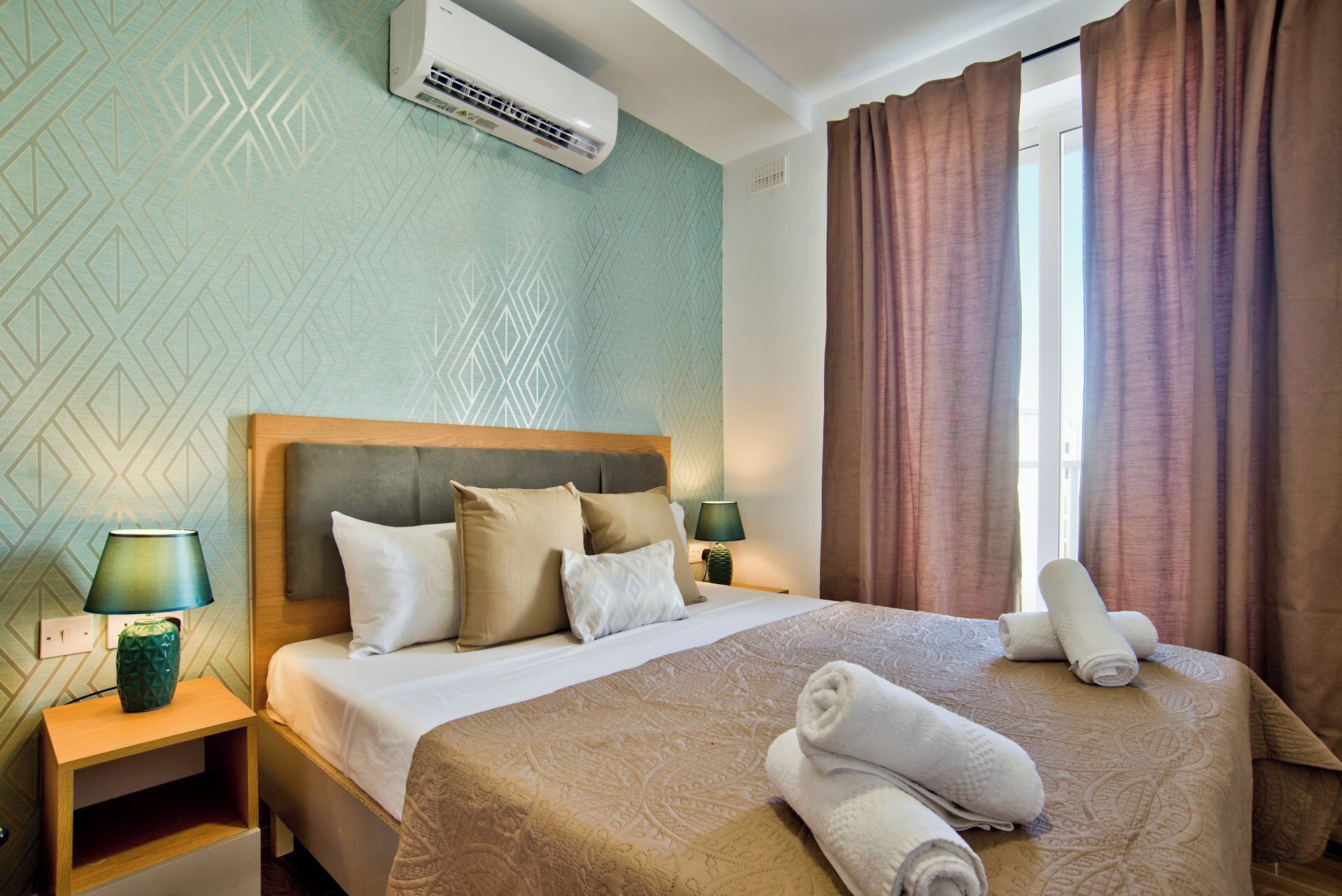 Ein Hotelzimmer mit einem Bett, zwei Nachttischen, einer Lampe, einem Fenster mit Vorhängen und einer Klimaanlage.