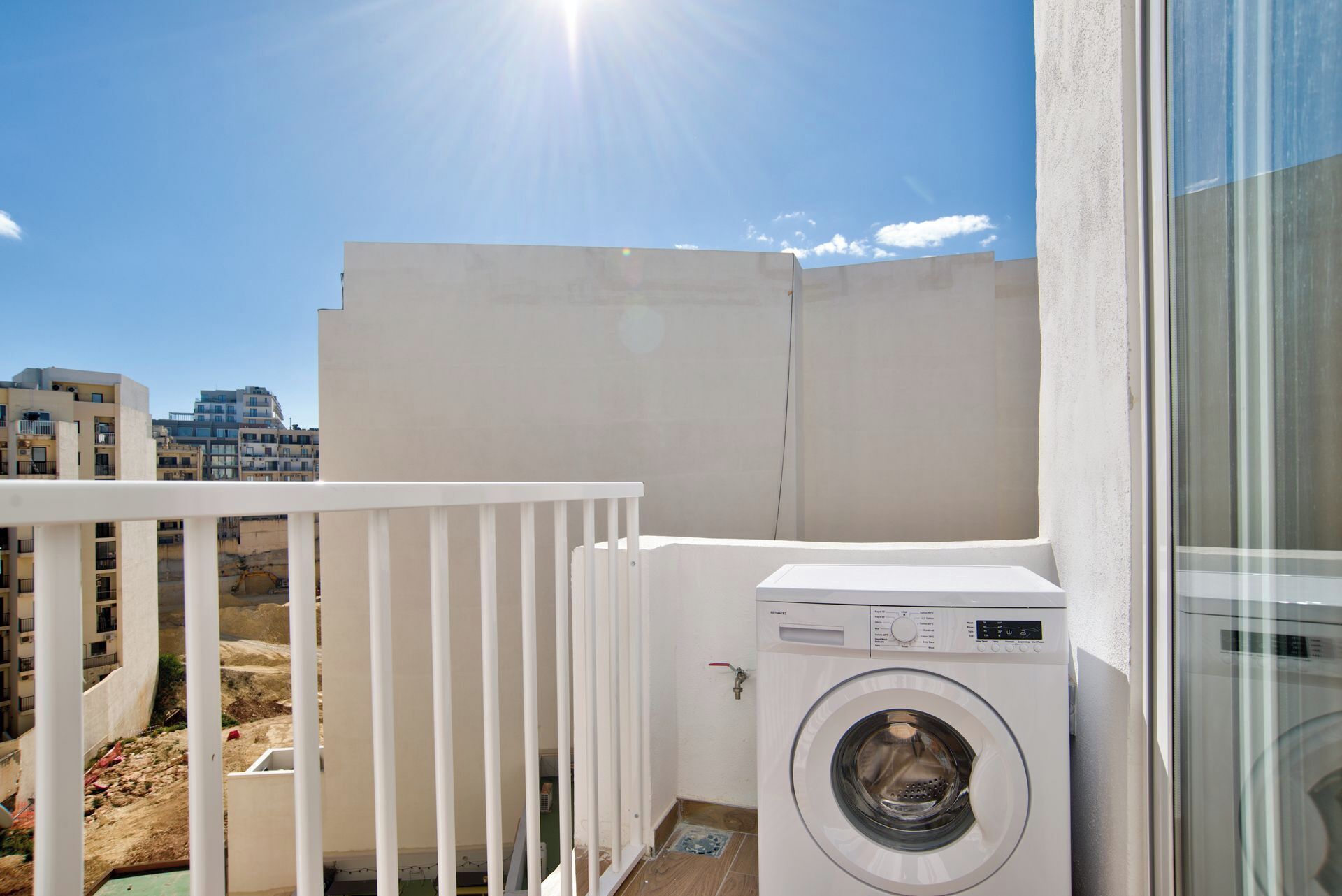 A balcony with a washing machine, a white railing, and a view of buildings in the background.