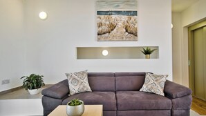 Uma sala de estar moderna com um sofá marrom escuro, almofadas estampadas, uma mesa de centro com uma planta, uma pintura de uma praia e uma pequena planta em um vaso dourado em uma estante.