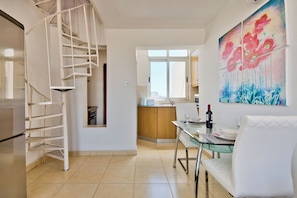 A modern kitchen with a spiral staircase, a dining area with a glass table and white chairs, and a vibrant floral painting on the wall. - Stunning City Views Sliema Duplex Penthouse (Sliema)