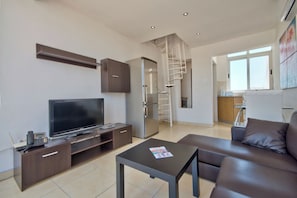 A modern living room with a flat-screen TV, a coffee table, a sofa, and a kitchen in the background. - Stunning City Views Sliema Duplex Penthouse (Sliema)