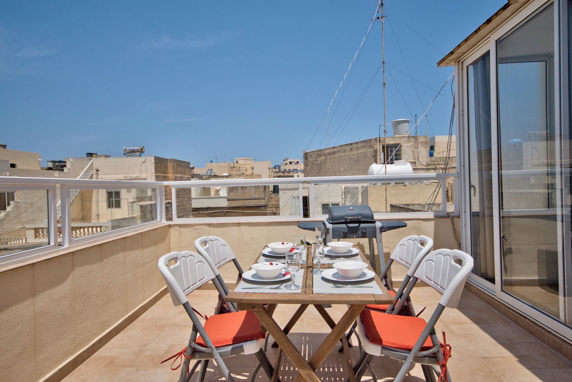 A rooftop terrace with a table set for four, a grill, and a view of the cityscape.