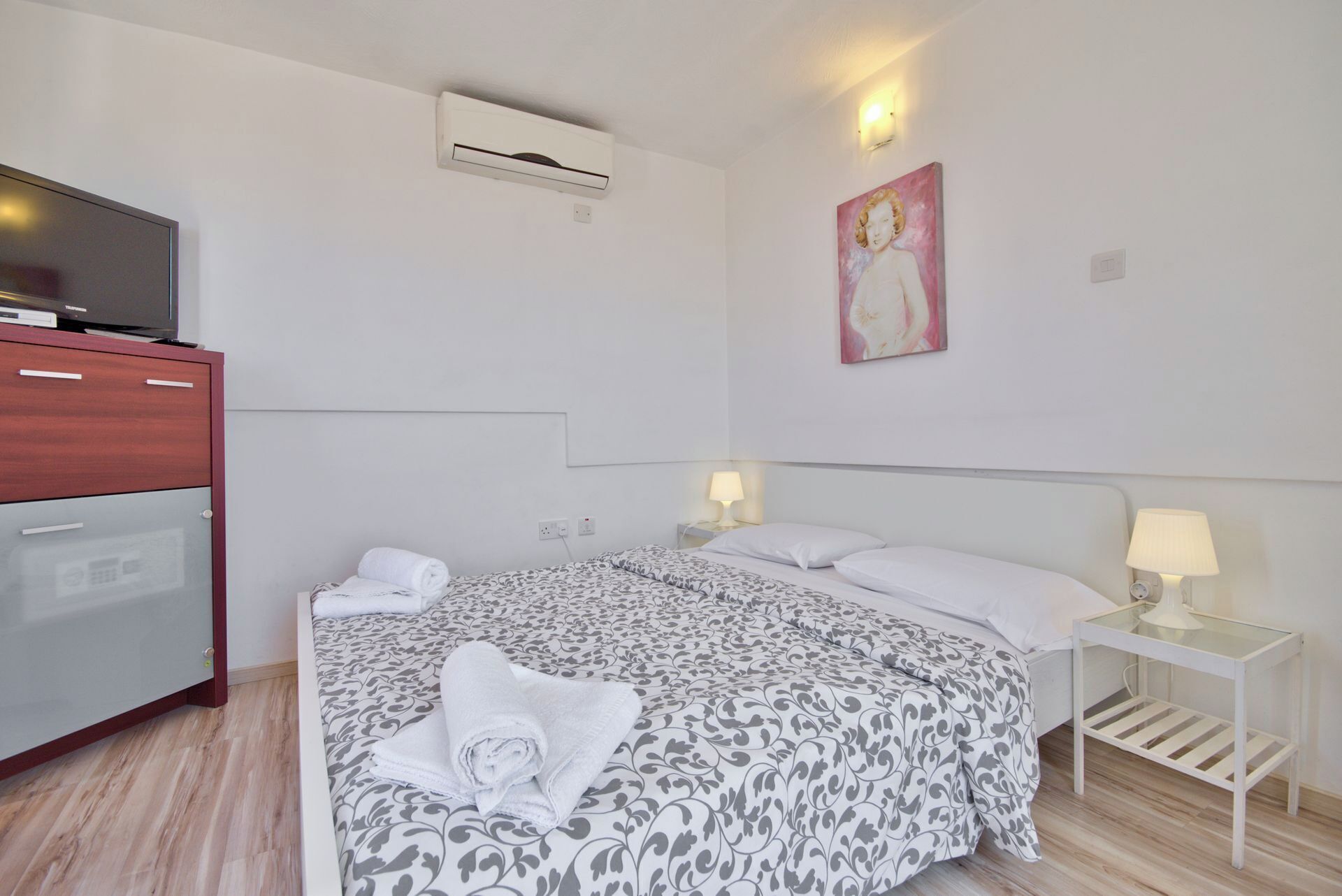 Een hotelkamer met een bed, nachtkastjes met lampen, een televisie, een kledingkast en een airconditioning.