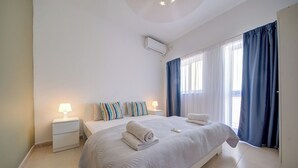 A bedroom with a large bed, two bedside tables, a window with curtains, and an air conditioning unit. - Off The Strand Gzira Top Floor Apartment (Gzira)