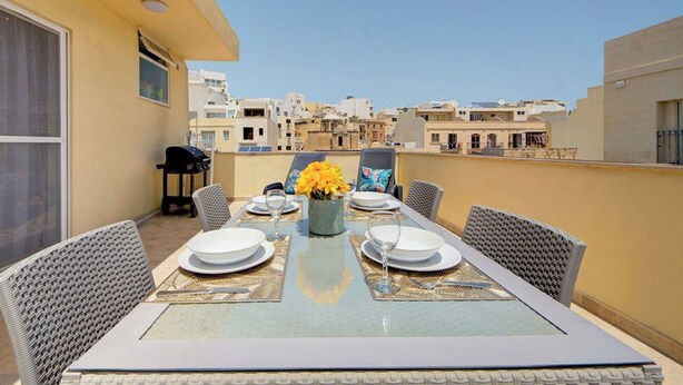 A rooftop terrace with a table set for dining, surrounded by wicker chairs, and a view of the cityscape. - Off The Strand Gzira Top Floor Apartment (Gzira)