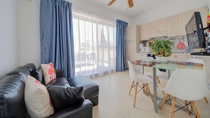 A modern living room with a black leather sofa, a dining table with chairs, a ceiling fan, and a kitchen area with wooden cabinets and a plant. - Off The Strand Gzira Top Floor Apartment (Gzira)