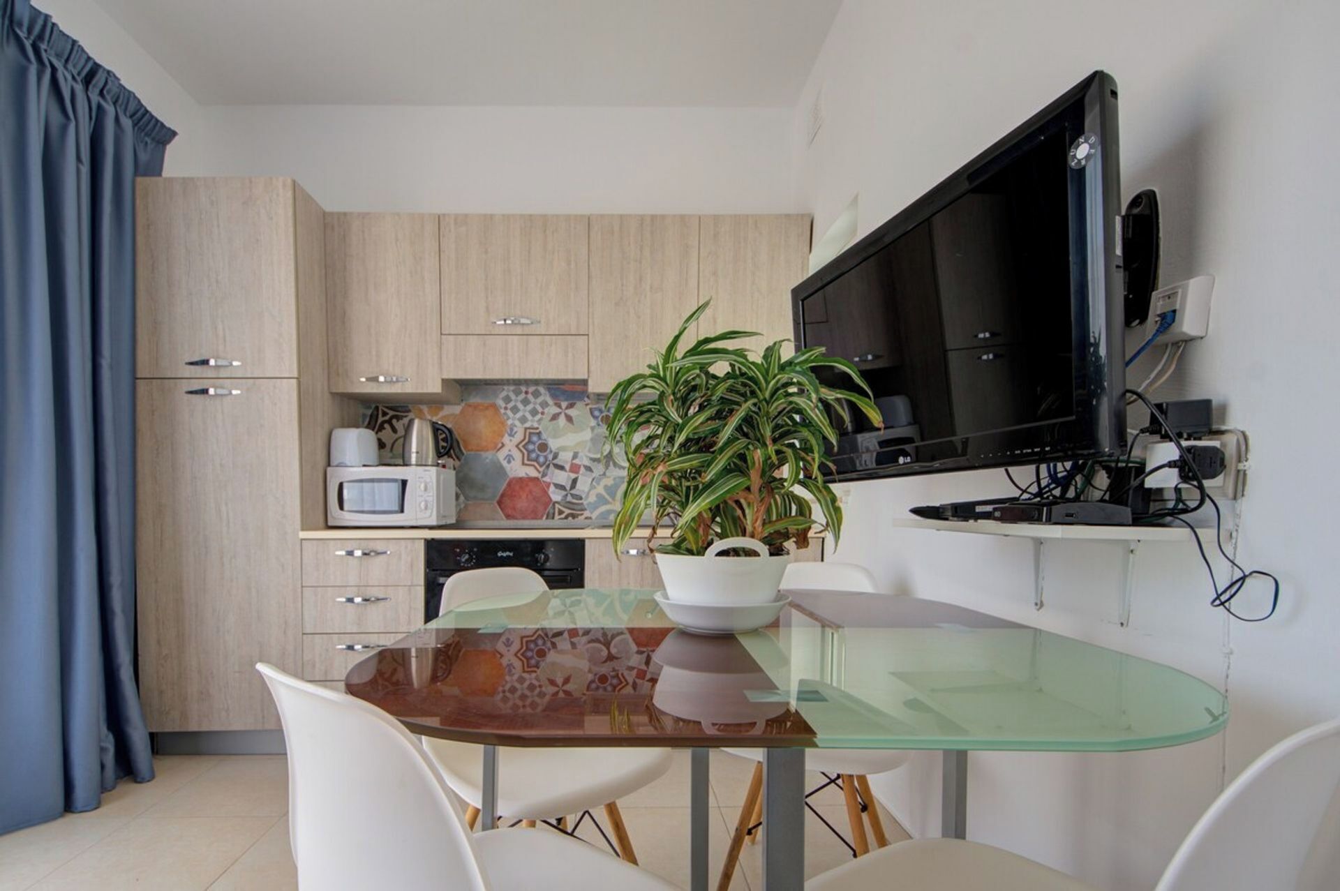 Een functionele keuken met een glazen eettafel, witte stoelen, een magnetron, koffiezetapparaat, een potplant en een aan de muur bevestigde televisie.