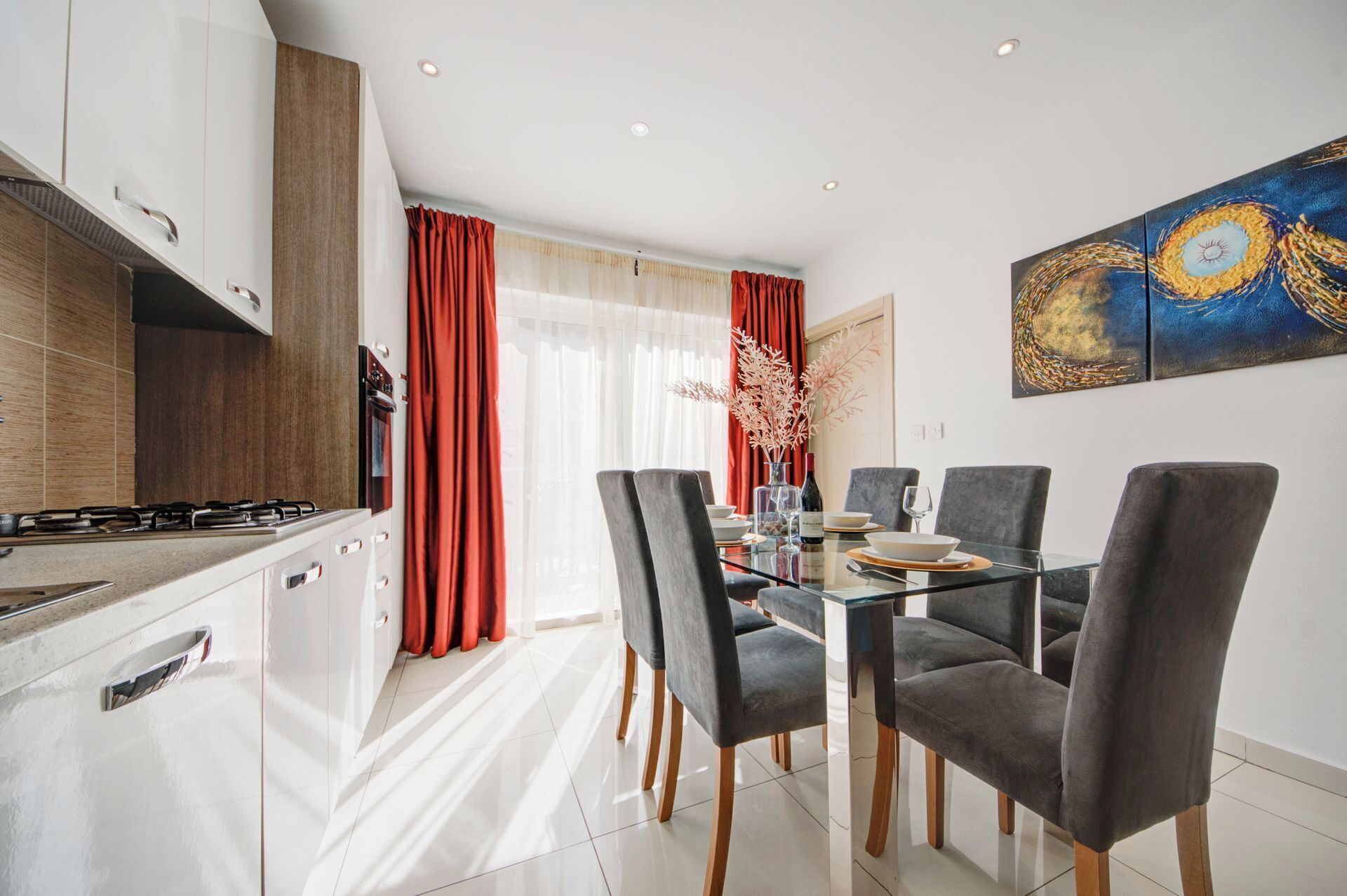 A modern kitchen with a gas stove, wooden cabinets, and a dining area with a glass table and grey upholstered chairs.