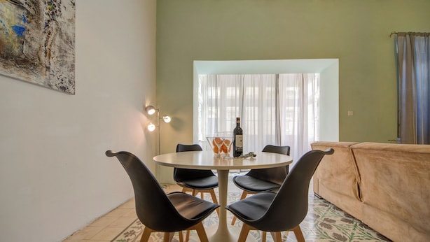 A dining area with a round table, four chairs, a bottle of wine, and a vase with oranges. - Spacious High Ceiling Sliema Apartment (Sliema)