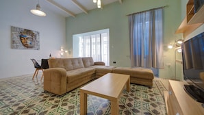 A living room with a sectional sofa, a small table, and a TV. There is a window with curtains, a painting on the wall, and a patterned floor. - Spacious High Ceiling Sliema Apartment (Sliema)