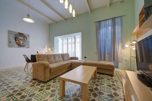 Un salon avec un canapé d’angle, une petite table et un téléviseur. Il y a une fenêtre avec des rideaux, un tableau au mur et un sol à motifs.