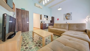 A living room with a sofa, a coffee table, and a TV. There is a kitchen area with cabinets and a microwave. The room has a light blue wall and a patterned floor. - Spacious High Ceiling Sliema Apartment (Sliema)
