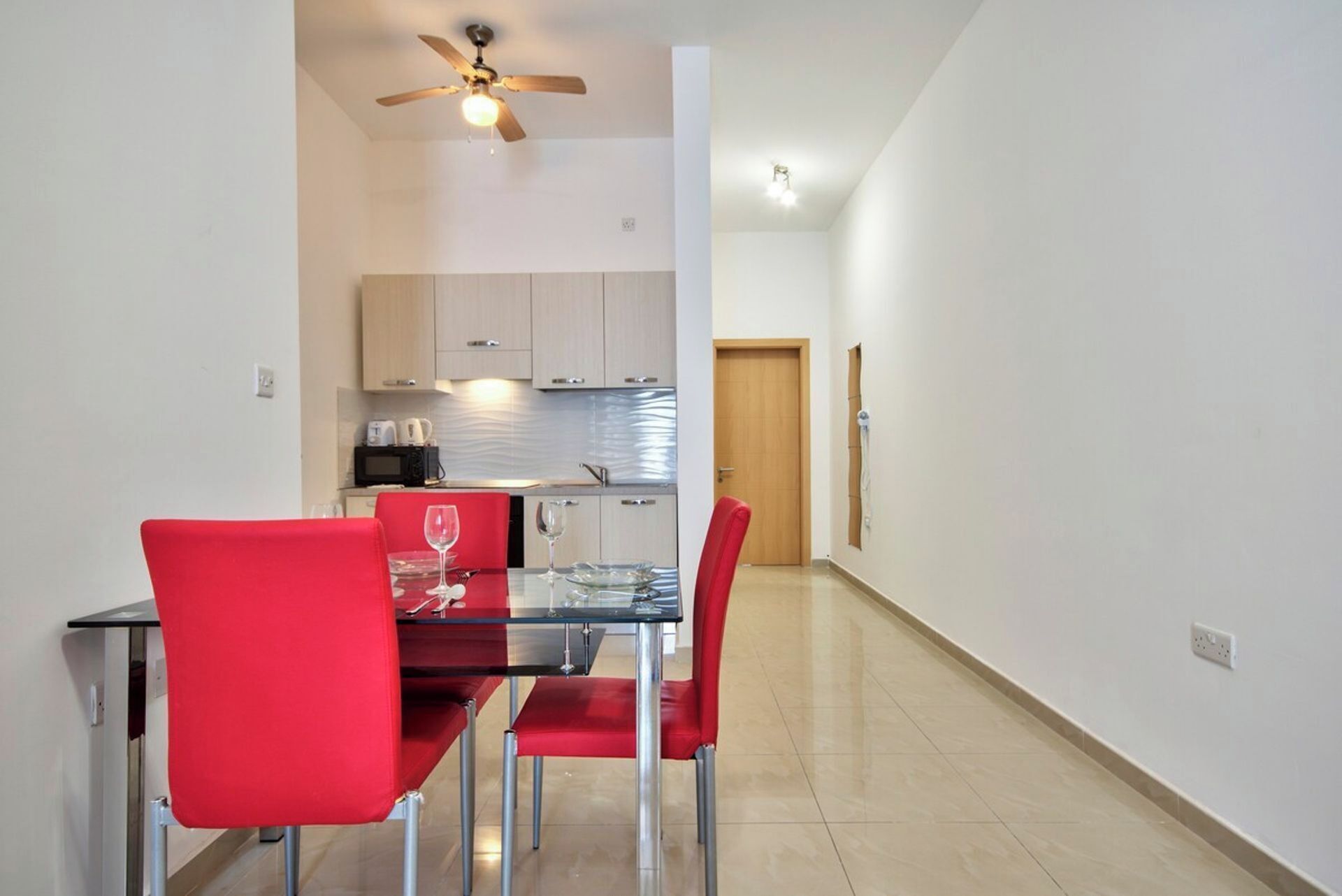 Una cocina moderna con armarios blancos, un microondas y un lavamanos. Hay una zona de comedor con una mesa de vidrio y sillas rojas.