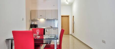 Una cocina moderna con armarios blancos, un microondas y un lavamanos. Hay una zona de comedor con una mesa de vidrio y sillas rojas.