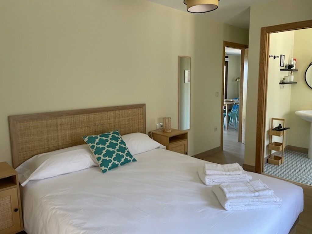 A neatly made bed with white linens and a teal pillow, a wooden nightstand with a lamp, and a bathroom visible through an open door.