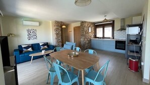 A modern living room with a blue sofa, a dining table with chairs, a kitchen area with cabinets and a stove, and a stone accent wall. - Apartment with shared swimming pool, Sta. (Baiona)
