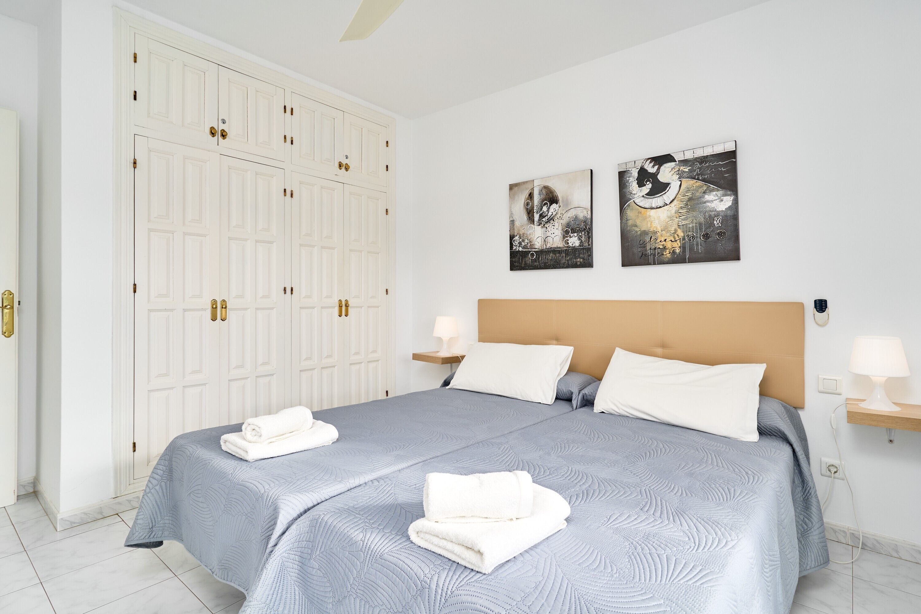 Un dormitorio con un armario grande, dos mesitas de noche con lámparas, una cama con cabecera y obras de arte en la pared.