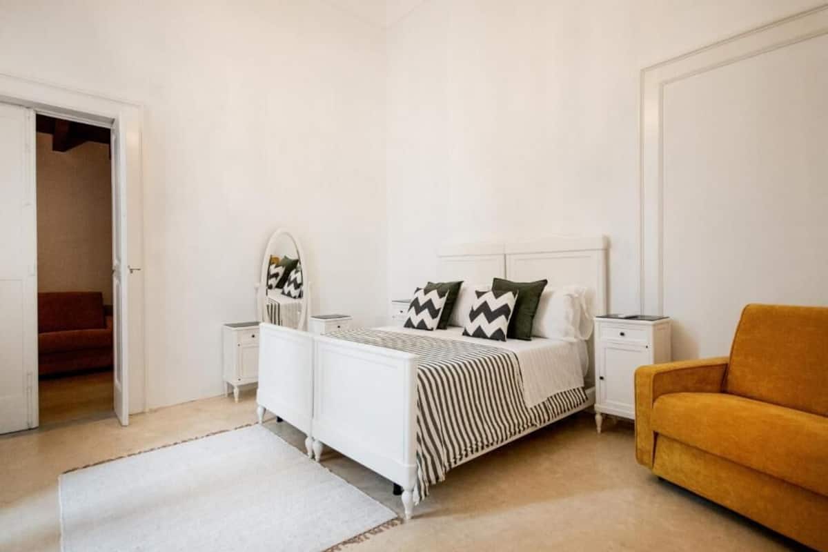 Een slaapkamer met een bed, nachtkastjes, een spiegel, een bank en een deur naar een andere kamer.