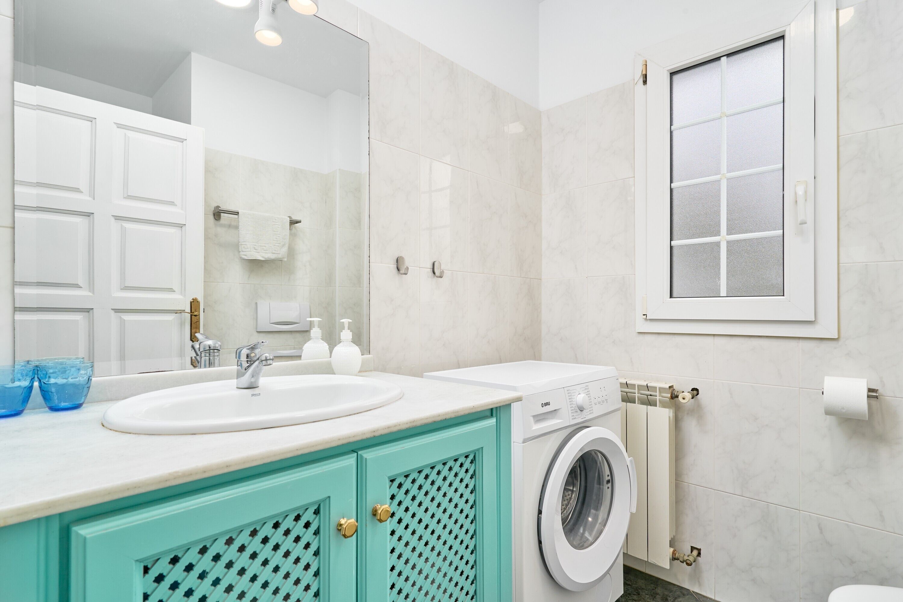 Un baño con un lavamanos blanco, una lavadora, un perchero para toallas y una ventana con vidrio esmerilado.