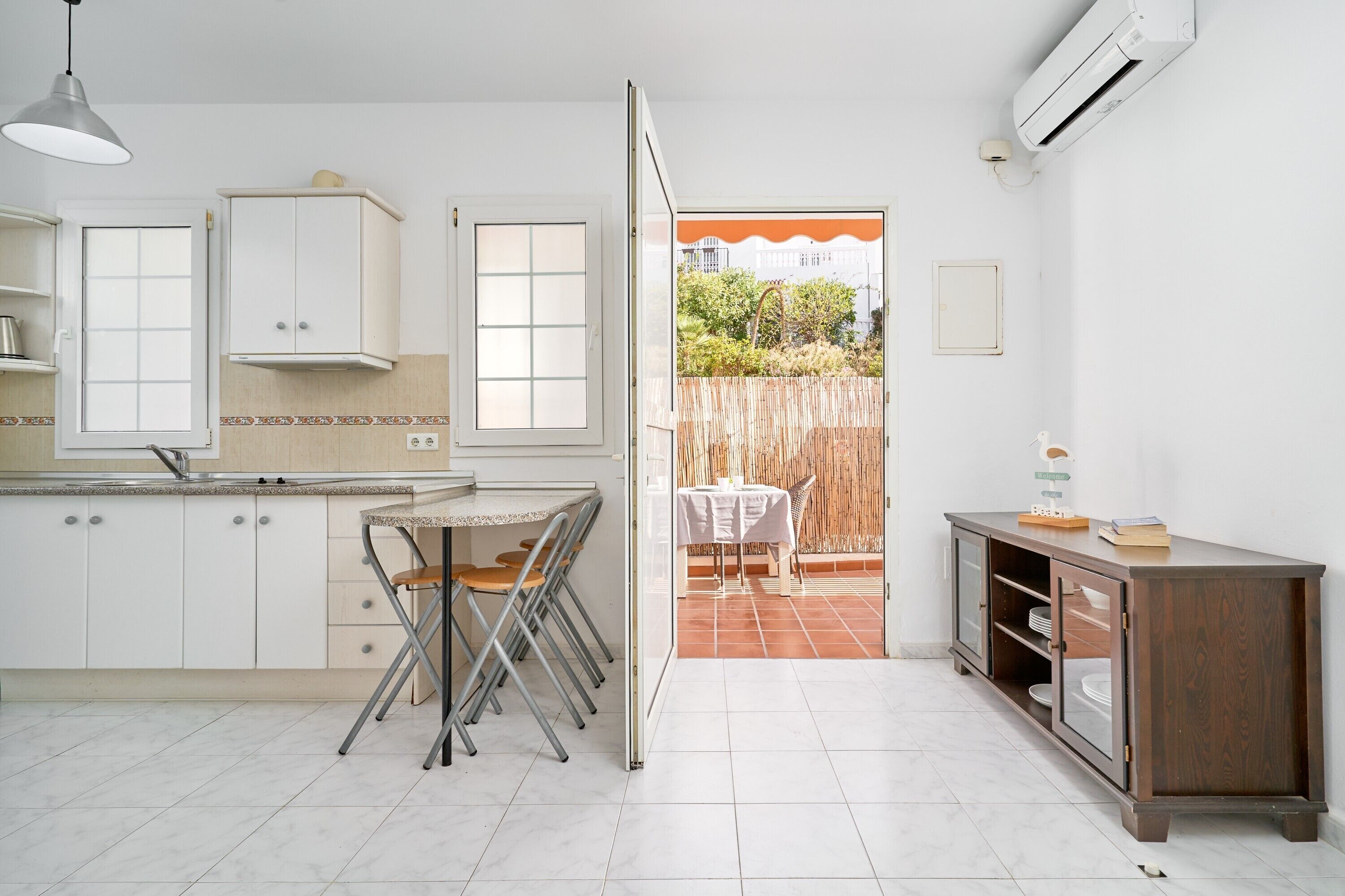 Une cuisine avec des meubles blancs, une table à manger avec des chaises, une porte ouverte donnant sur un patio et un appareil de climatisation.