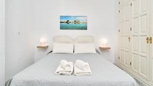 A neatly made bed with two pillows, a pair of towels, a painting of a beach scene, and a wardrobe with brass handles. - Apartment in A Hotel 'B-B' with Shared Pool, Wi-Fi and Air Conditioning (Nerja)