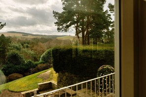 Blick aus einem Fenster auf einen angelegten Garten mit Steinmauer, Rasenfläche und Bäumen.