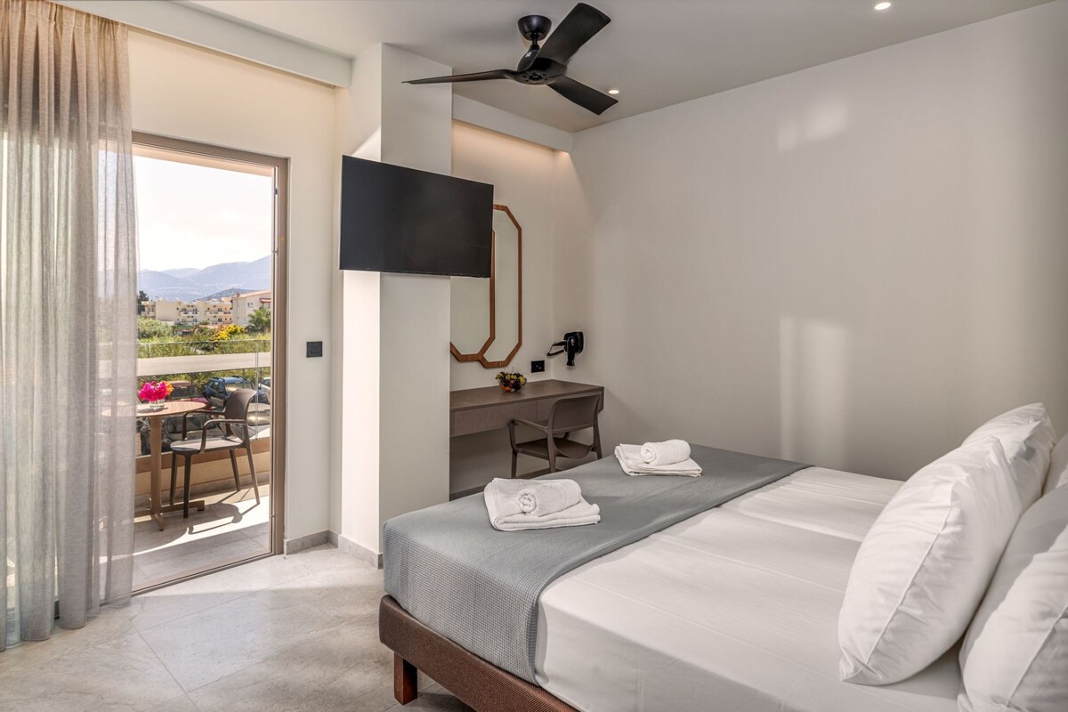 Habitación de hotel con cama, escritorio, silla, televisor y balcón con vistas a edificios y montañas.