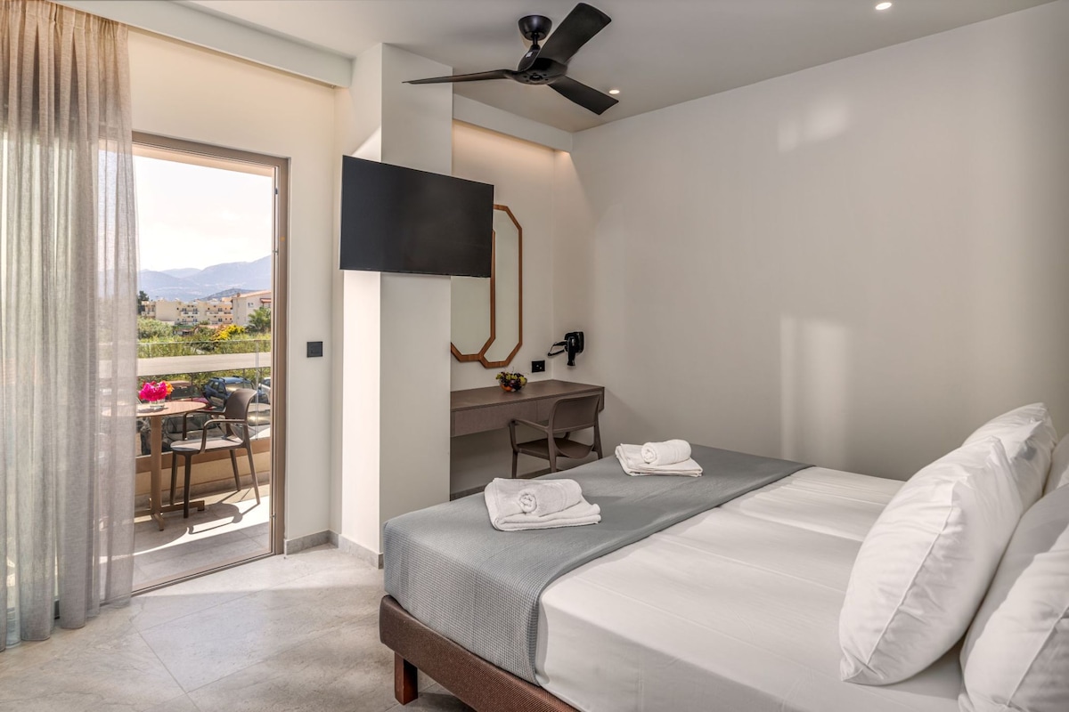 Una camera d'albergo con un letto, una scrivania, una sedia, una TV e un balcone con vista su edifici e montagne.