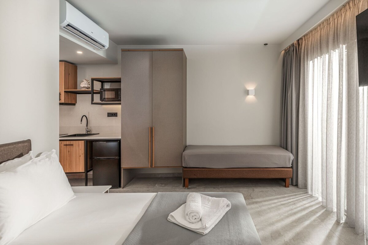 Habitación de hotel con cama, escritorio con lavamanos, armario y ventana grande con cortinas.