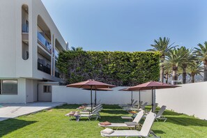 A poolside area with lounge chairs and umbrellas, a hedge, and a building with balconies. - Malia Odyssey Adults Only by Estia (Malia)