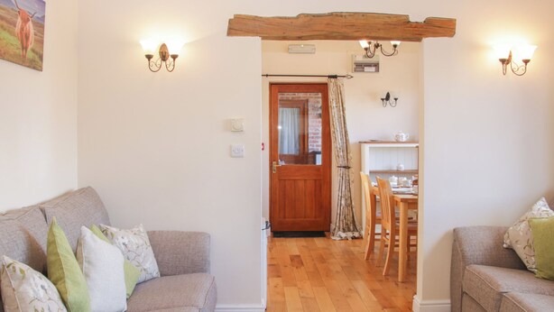 A cozy living room with a wooden floor, two sofas with cushions, a dining table with chairs, and a kitchen area with a wooden door and light fixtures. - Herdwick Cottage (Much Wenlock)