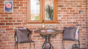 A brick wall with a wooden-framed window, a self-catering sign, and two metal chairs with patterned cushions. - Herdwick Cottage (Much Wenlock)