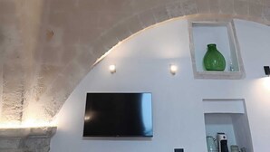 A room with a stone ceiling, a flat-screen TV mounted on the wall, and a small shelf with a green vase. - At Vico 12 (Monopoli)