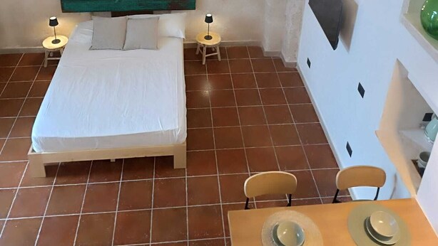 A bedroom with a bed, two bedside tables with lamps, a wooden headboard, a small table with chairs, and a tiled floor. - At Vico 12 (Monopoli)