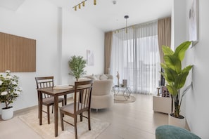A modern living room with a dining area, a sofa, and a coffee table. There are potted plants and a large window with curtains. - Luxurious JVC Getaway l Infinity Pool & Gym (Dubai)