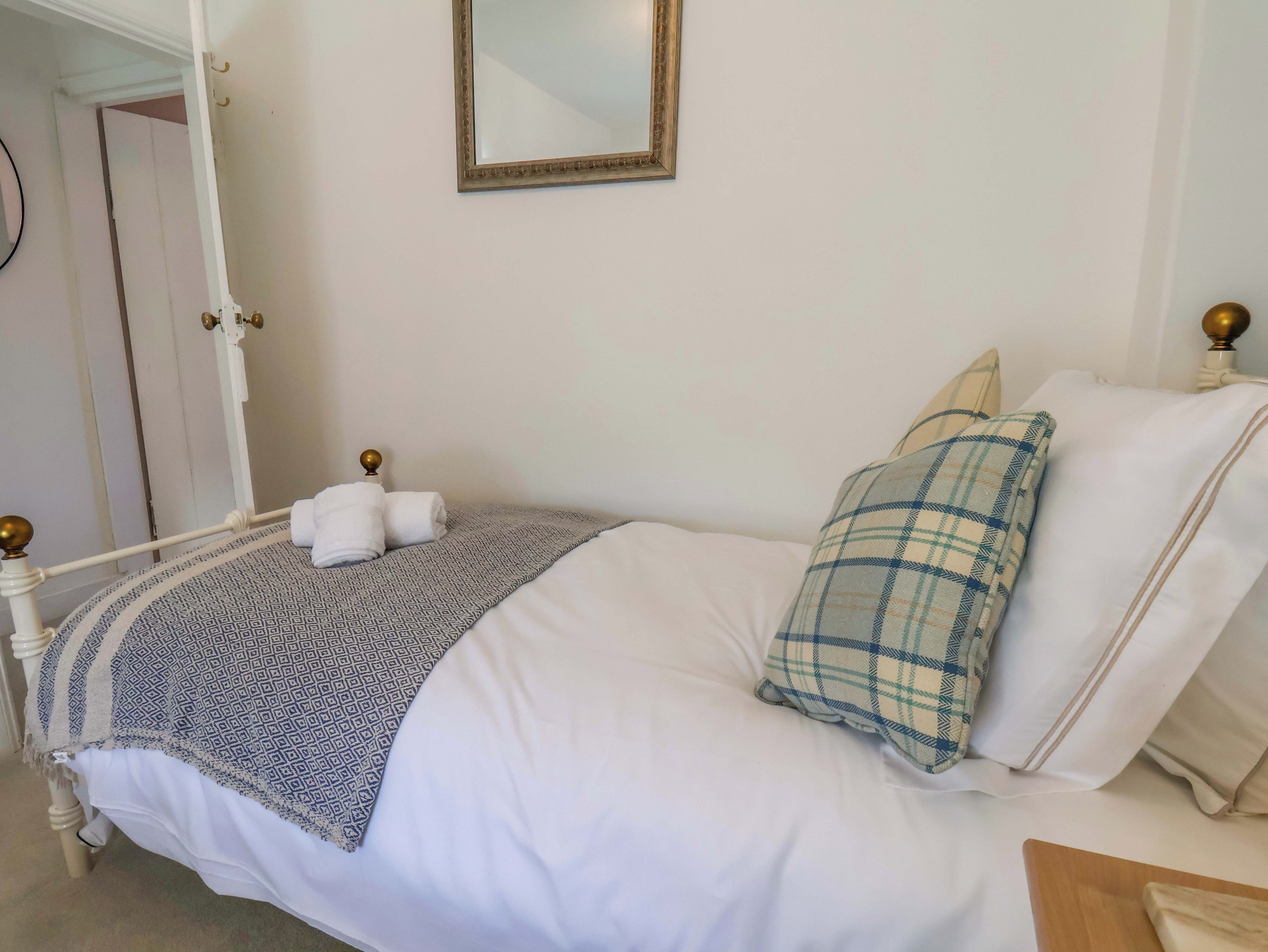 A neatly made bed with a patterned blanket, a plaid pillow, and a white pillow. There is a small bedside table with a lamp and a mirror on the wall above the bed.