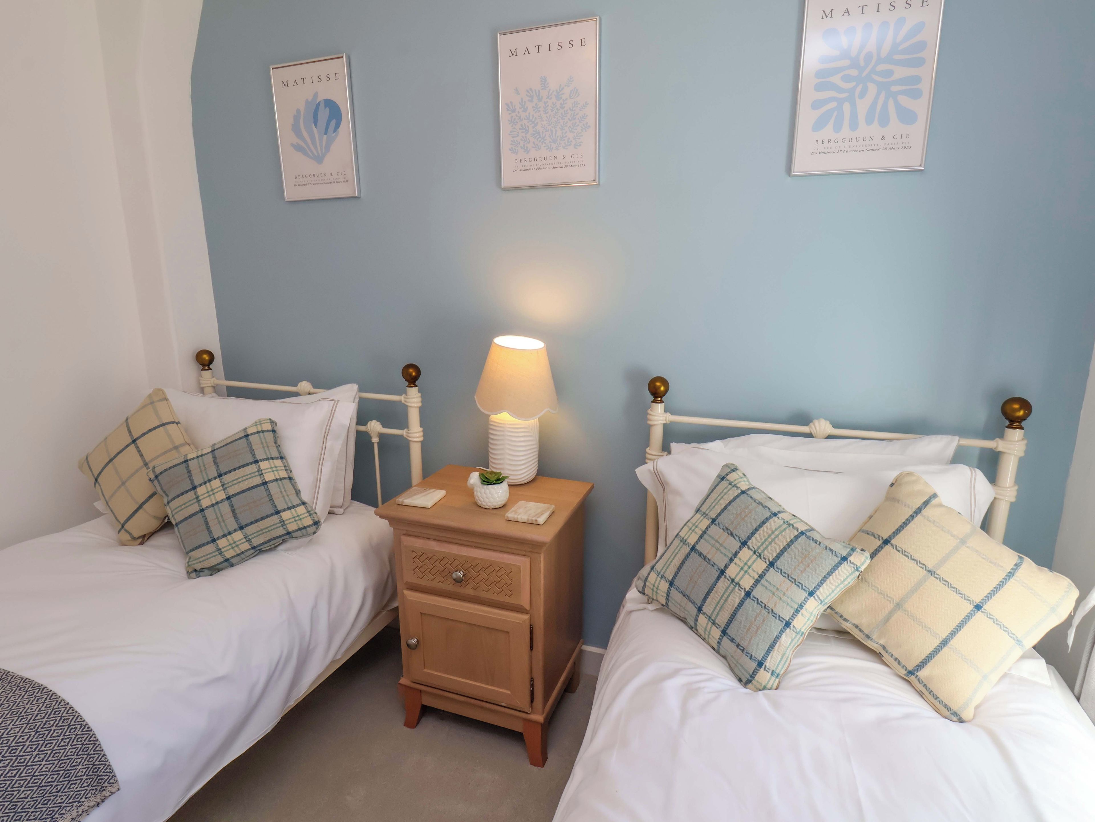 A bedroom with two single beds, a nightstand with a lamp, and three framed artworks on the wall.