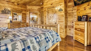 Interior de una cabaña de madera con una cama, cómoda y televisor montado en la pared.