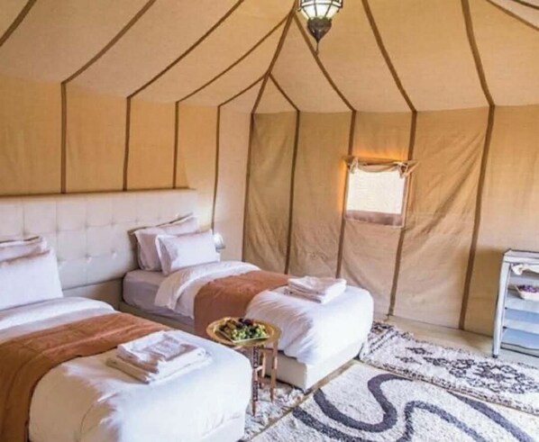 A tent-style room with two beds, a small table with a fruit bowl, and a window. - Majestic Luxury Tented  (Merzouga)