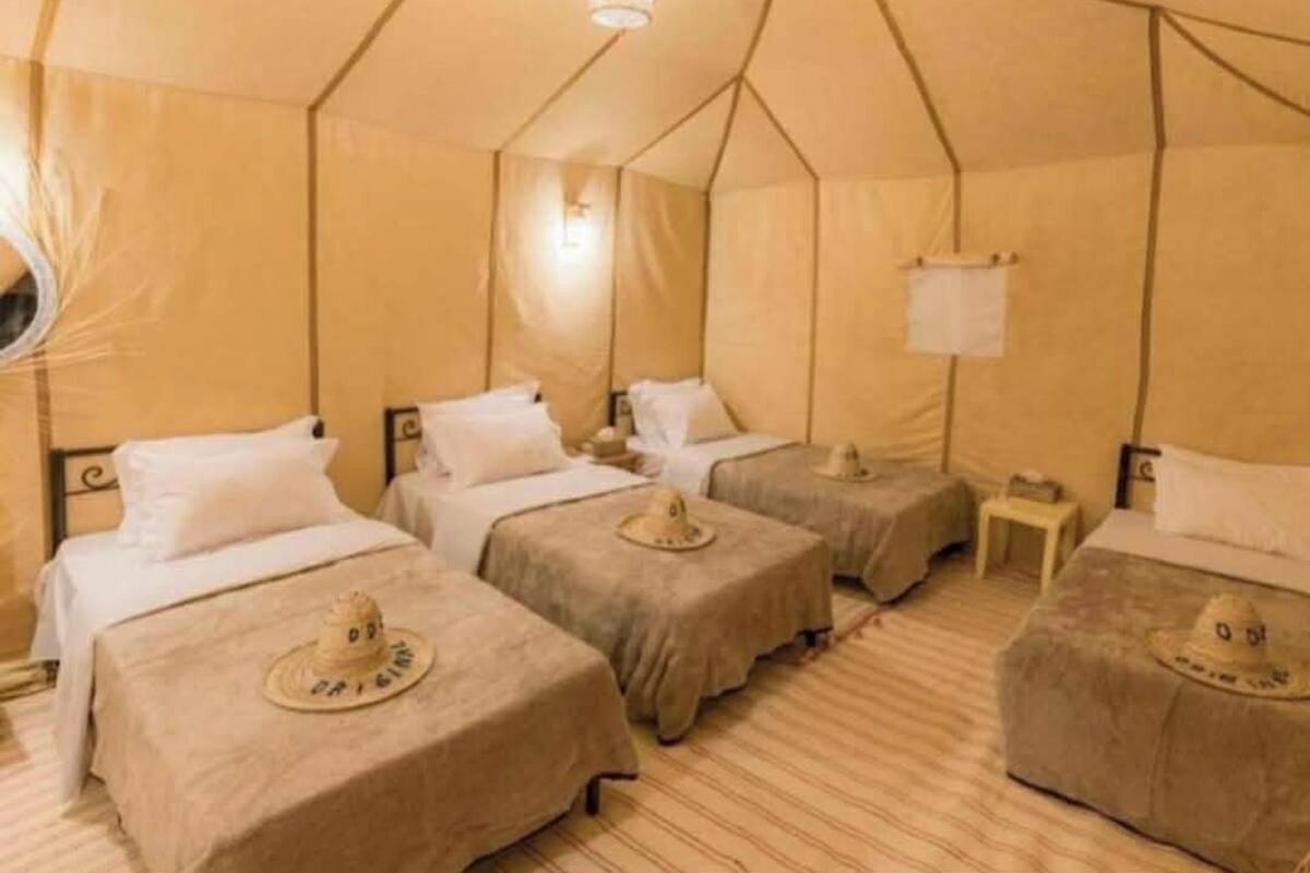 Une pièce aménagée comme une tente, avec quatre lits, chacun surmonté d’un couvre-lit orné d’un chapeau, une petite table et un luminaire suspendu.