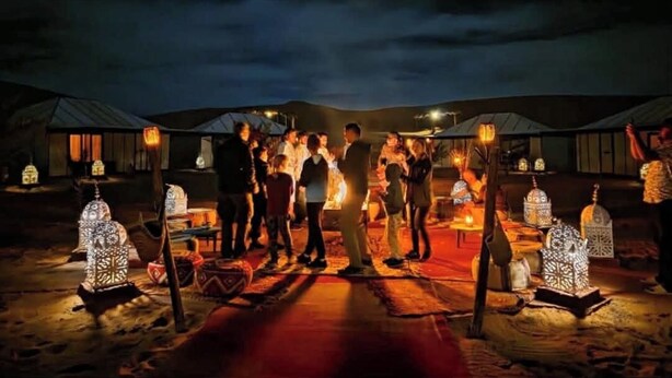 A group of people gathered around a central fire in a desert setting with tents in the background. - Majestic Luxury Tented  (Merzouga)