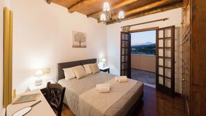 Ein Schlafzimmer mit einem Bett, Nachttischen, einem Schreibtisch, einem Stuhl, einer Chandelier und Blick durch eine offene Tür.