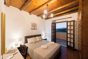 A bedroom with a bed, bedside tables, a desk, a chair, a chandelier, and a view through an open door. - Abrakamus by Forest Villas (Rethymno)